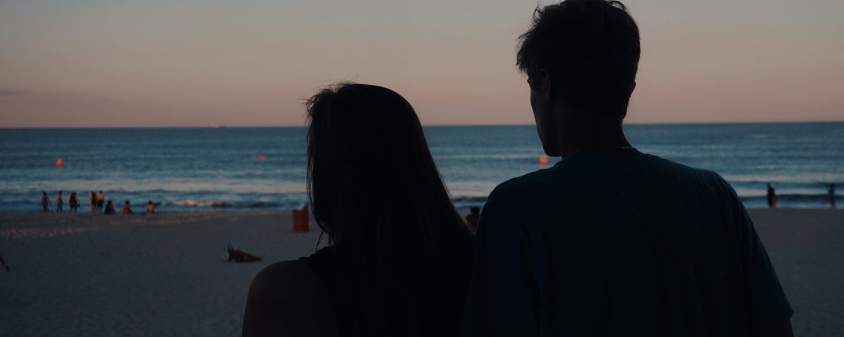 A man and woman watch the sunset on the beach.