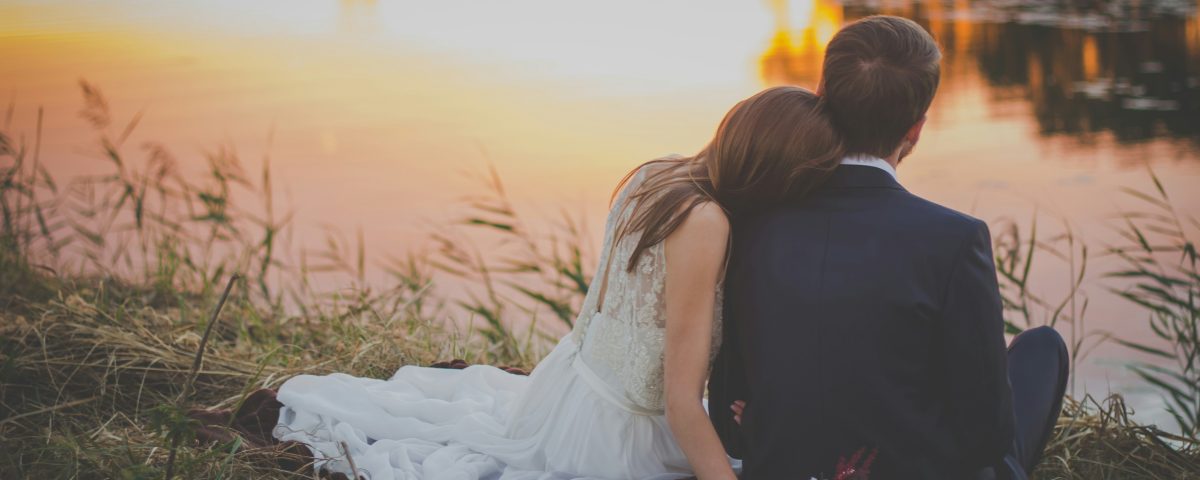 A newlywed couple sit by a lake.