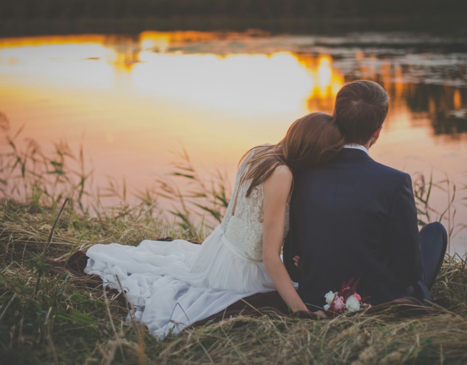 A newlywed couple sit by a lake.